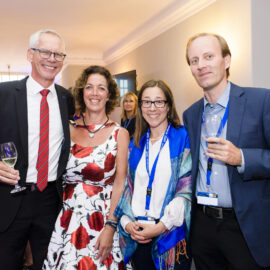 Vier Personen stehen im Vordergrund einer festlichen Veranstaltung, alle in formeller Kleidung. Zwei Männer tragen Anzüge und halten jeweils ein Glas Weißwein, während die beiden Frauen elegante Kleider tragen, eine davon mit einem blauen Schal und Namensschild. Im Hintergrund sind weitere Gäste in festlicher Kleidung im Gespräch.