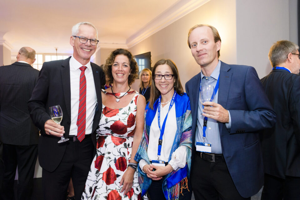 Vier Personen stehen im Vordergrund einer festlichen Veranstaltung, alle in formeller Kleidung. Zwei Männer tragen Anzüge und halten jeweils ein Glas Weißwein, während die beiden Frauen elegante Kleider tragen, eine davon mit einem blauen Schal und Namensschild. Im Hintergrund sind weitere Gäste in festlicher Kleidung im Gespräch.