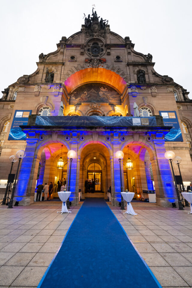 Der Eingang eines prunkvollen Gebäudes ist in blauem und gelbem Licht beleuchtet. Die Architektur zeigt große Rundbögen, verzierte Details und Statuen. Ein blauer Teppich führt zum Eingang, gesäumt von kleinen runden Tischen mit weißen Tischdecken.
