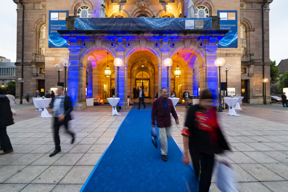 Der Eingang eines eindrucksvollen Gebäudes ist in blauem und violettem Licht angestrahlt, wodurch Bögen und Säulen hervorgehoben werden. Ein blauer Teppich führt zum Eingang, von dem aus mehrere Personen unterwegs sind. Vor dem Gebäude stehen runde Tische mit weißen Tischdecken.