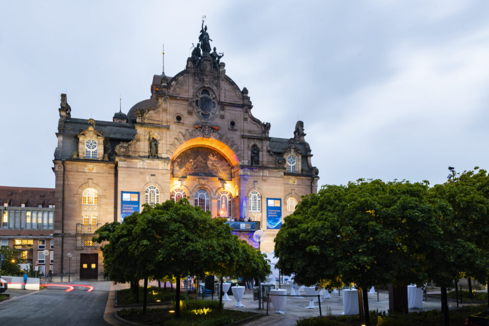 Ein historisches Gebäude mit aufwendiger Architektur und großem Rundbogen-Eingang ist beleuchtet und hebt so seine Details hervor. Davor befinden sich mehrere Bäume sowie ein Außenbereich mit Tischen und Stühlen, was auf eine Veranstaltung hinweist. Am Gebäude sind zudem Banner angebracht, der Himmel ist bewölkt.