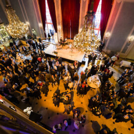 Ein großer, eleganter Saal mit hohen Decken und prunkvollen Kronleuchtern ist mit vielen Menschen gefüllt. Einige Gäste stehen in Gruppen, andere sitzen an runden Tischen. Auf einer Bühne sind Musikinstrumente aufgebaut, was auf eine bevorstehende Aufführung oder Veranstaltung hinweist.