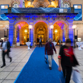 Der Eingang eines eindrucksvollen Gebäudes ist in blauem und violettem Licht angestrahlt, wodurch Bögen und Säulen hervorgehoben werden. Ein blauer Teppich führt zum Eingang, von dem aus mehrere Personen unterwegs sind. Vor dem Gebäude stehen runde Tische mit weißen Tischdecken.