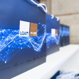 Mehrere blaue Geschenktaschen mit weißen Kordelgriffen stehen auf einem weißen Regal. Die Taschen zeigen ein Muster aus verbundenen Linien und Punkten.