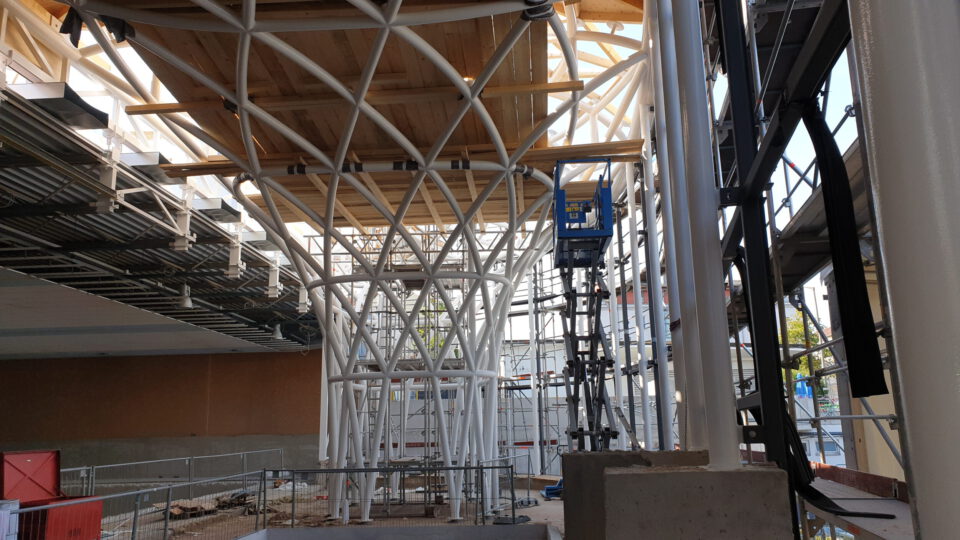 Baustelle mit komplexem Stahlgerüst, das eine Holzkonstruktion trägt; umgeben von Gerüsten, Leitern und einem blauen Hubsteiger, der Bereich ist mit Metallzäunen abgesperrt und mit Baumaterialien übersät.