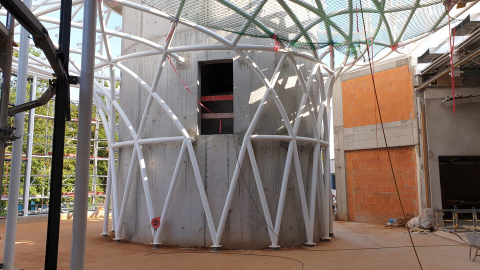 Baustelle mit zylindrischer Betonstruktur im Zentrum, umgeben von einem weißen Metallgerüst aus diagonalen und vertikalen Trägern; der Boden ist sandig, mit verstreuten Baumaterialien, im Hintergrund ein weiteres Gebäude mit sichtbarem Ziegelmauerwerk.