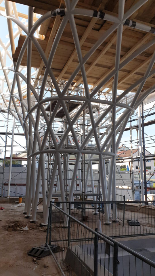 Baustelle mit komplexem Gerüst aus weißen, sich kreuzenden Metallträgern, die eine hölzerne Plattform tragen; umgeben von Gerüsten, Sicherheitsgeländern und Baumaterialien, im Hintergrund weitere Gebäude und klarer Himmel.