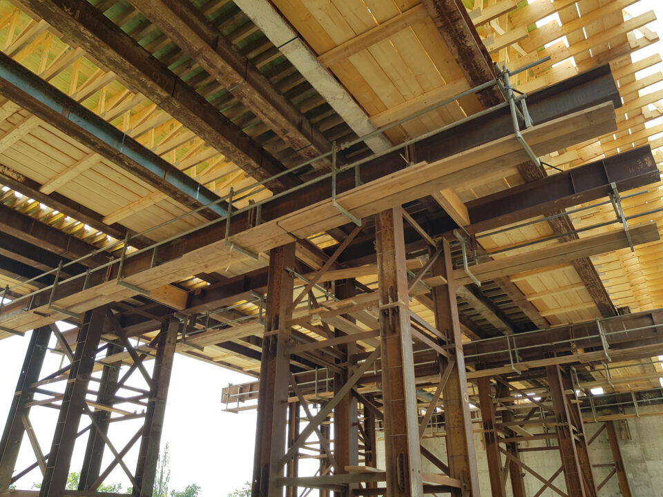 Blick auf die Unterseite einer Baustelle mit Gerüsten und Trägern, die eine hölzerne Deckenkonstruktion stützen; sichtbar sind Metallstützen, Holzbalken und Planken, die das obere Stockwerk tragen.