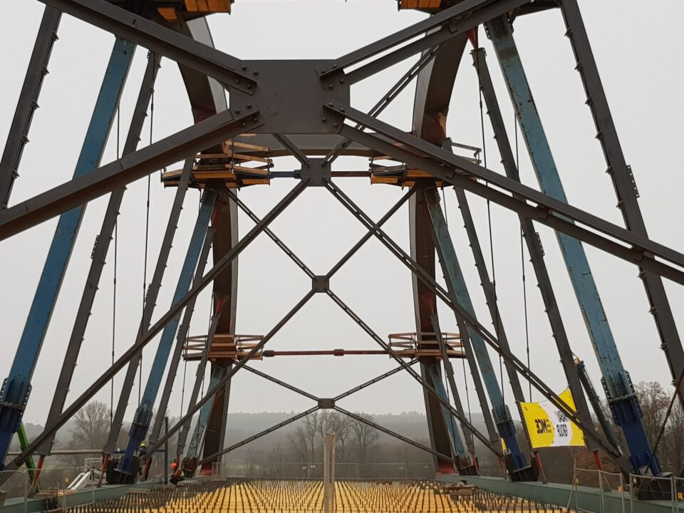 Nahaufnahme einer Stahlbrücke im Bau mit großen Stahlträgern in dreieckiger Anordnung sowie zusätzlichen Stützbalken und sichtbarem Gerüst.