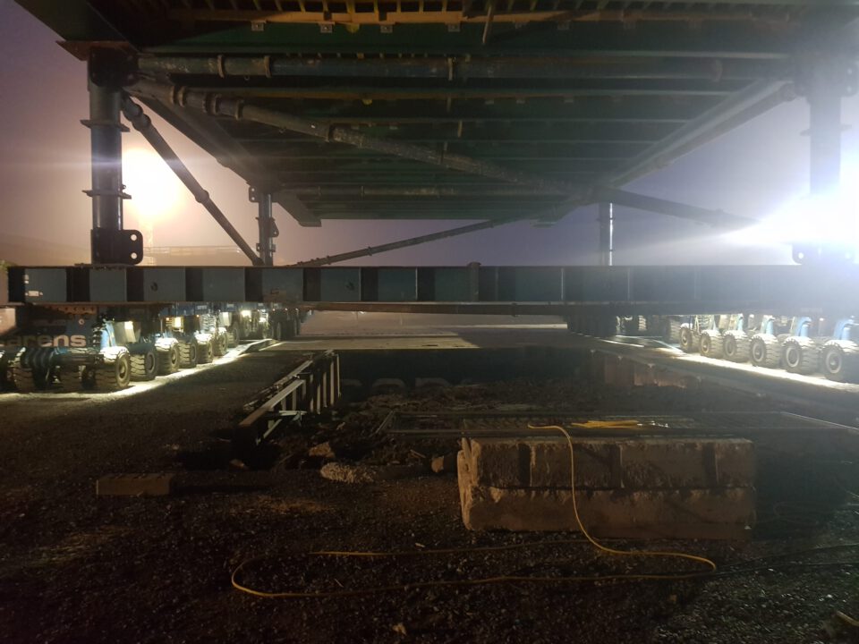 Große industrielle Struktur, die von mehreren Radachsen getragen und bewegt wird; die Szene spielt bei Nacht oder in schwachem Licht, beleuchtet durch künstliche Lichtquellen.