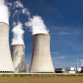 Das Bild zeigt ein Kernkraftwerk mit vier großen Kühltürmen, aus denen Dampf aufsteigt. Im Vordergrund liegt ein offenes Feld mit etwas Vegetation, der Himmel ist blau mit vereinzelten Wolken.
