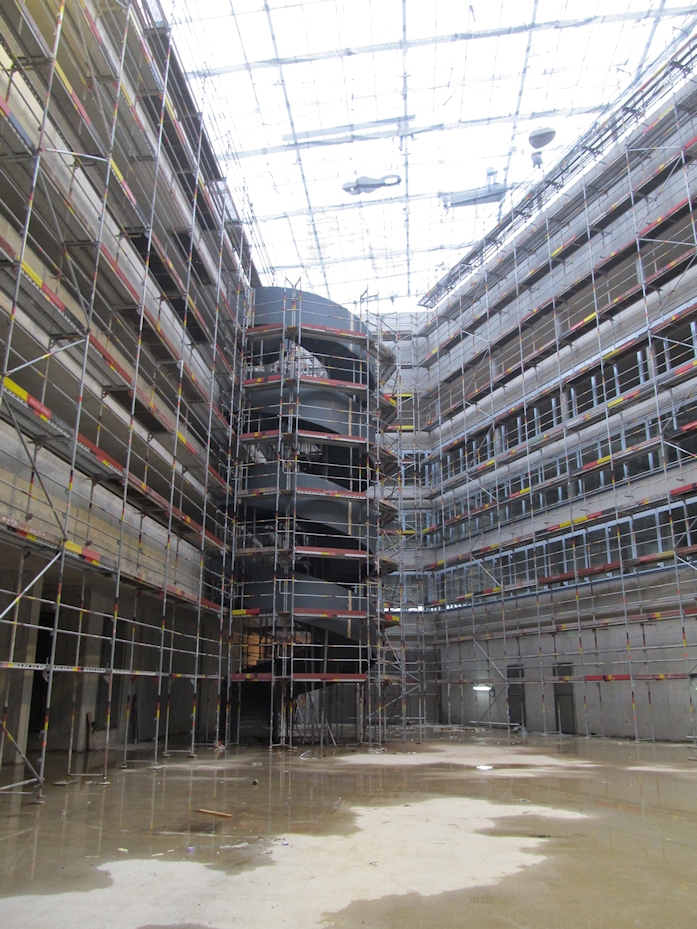 Das Innere eines großen Gebäudes im Bau mit mehrstöckigem Gerüst, einem teilweise fertiggestellten Treppenhaus oder Aufzugsschacht in der Mitte und einem nassen Boden; das provisorisch abgedeckte Dach lässt Licht hindurchscheinen.