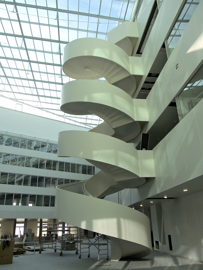 Ein modernes Atrium mit einer großen weißen Wendeltreppe, die sich über mehrere Etagen erstreckt; durch das Glasdach fällt Tageslicht in den hellen Raum mit weißen Wänden, Glasgeländern und sichtbaren Fensterfronten.