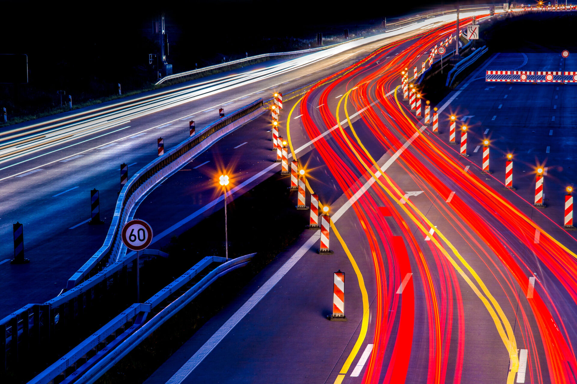 Das Bild zeigt eine nächtliche Autobahnszene mit roten und gelben Lichtspuren vorbeifahrender Fahrzeuge. Verkehrskegel, Absperrungen und ein Tempo-60-Schild wegen Bauarbeiten und Fahrbahnverengung.