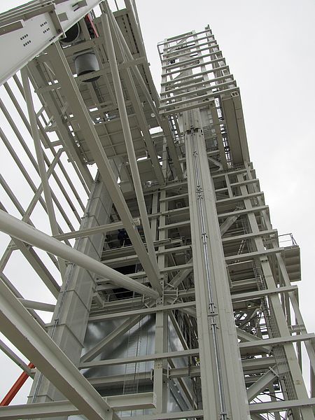 Das Bild zeigt eine Nahaufnahme eines großen, komplexen Industriebaus aus Metallträgern und Plattformen mit mehreren Ebenen, Leitern und Geländern, aus der Froschperspektive gegen einen wolkenverhangenen Himmel.