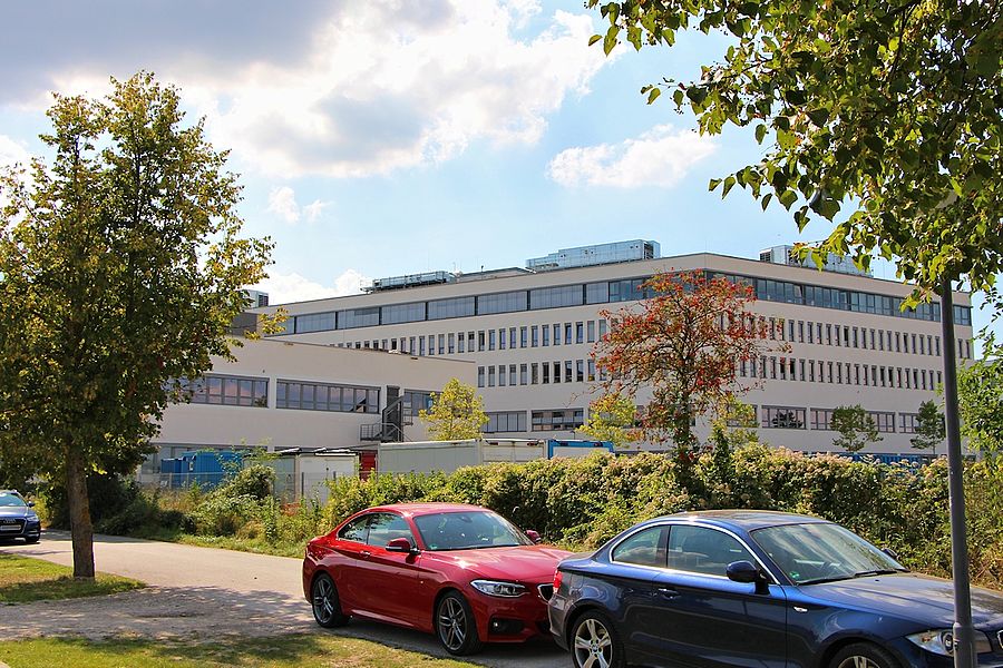Ein großes, modernes Gebäude mit mehreren Etagen und zahlreichen Fenstern steht im Hintergrund; im Vordergrund sind ein rotes und ein blaues Auto geparkt, umgeben von Bäumen, Grünflächen und einem teils bewölkten Himmel.