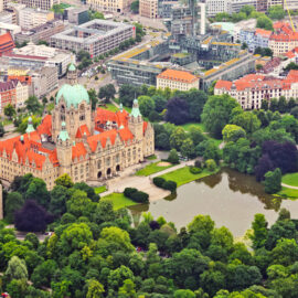 Luftaufnahme von Hannover mit dem Neuen Rathaus im Zentrum, erkennbar an seinem roten Dach und der grünen Kuppel, umgeben von Parkanlagen mit Teich und einer Mischung aus moderner und traditioneller Stadtarchitektur.