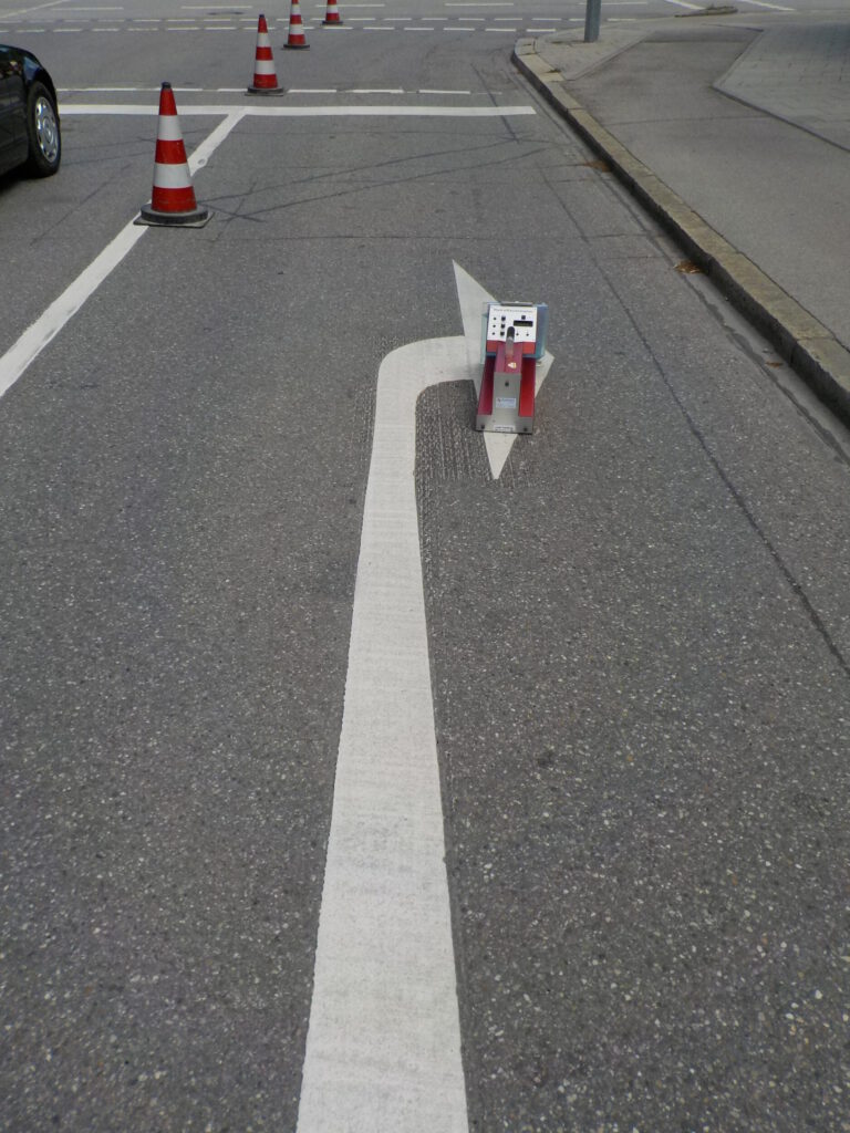 Das Bild zeigt einen Straßenabschnitt mit weißen Fahrbahnmarkierungen, darunter ein Pfeil, der eine Rechtsabbiegung anzeigt. Entlang der Fahrspur stehen mehrere orangefarbene Leitkegel, und eine kleine rot-weiße Absperrung befindet sich mittig auf dem Pfeil. Im Hintergrund sind weitere Fahrspuren, Gehwege und Bordsteine zu sehen.