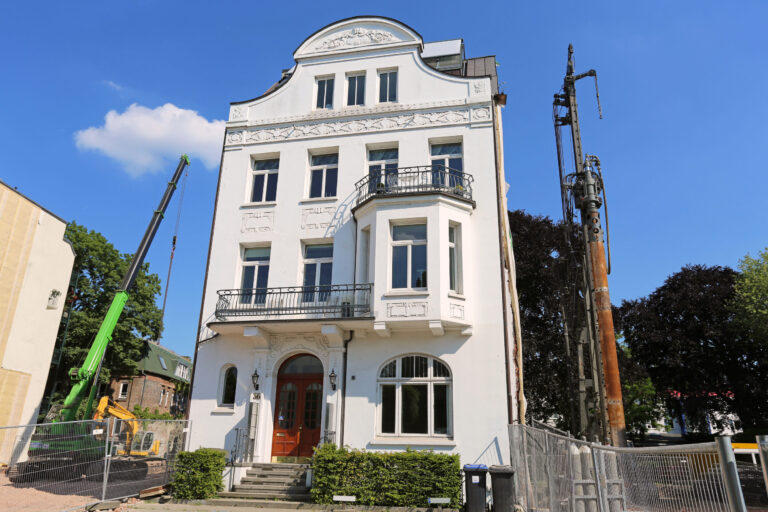 Das Bild zeigt ein weißes, mehrstöckiges Gebäude mit aufwendigen architektonischen Details. Mehrere Balkone mit schwarzen Geländern und große Fenster prägen die Fassade. Auf der linken Seite befinden sich Baumaschinen, darunter ein Kran und eine Bohrvorrichtung.