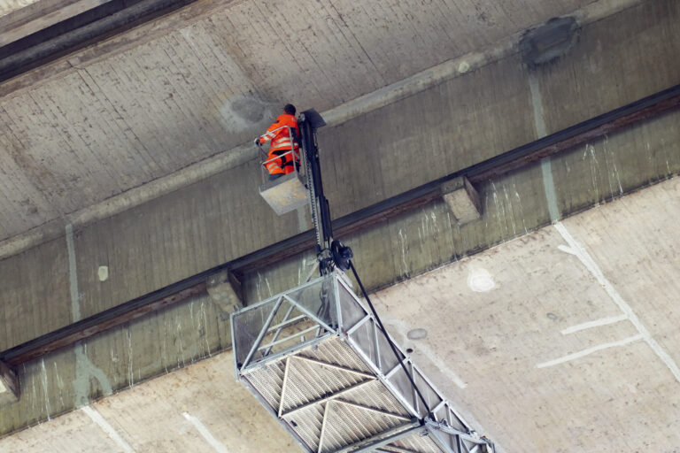 Das Bild zeigt eine Person in einem orangefarbenen Schutzanzug, die auf einer angehobenen Arbeitsplattform steht und an die Unterseite eines großen Betonbauwerks, einer Brücke inspiziert. Die Plattform ist an einem ausfahrbaren Arm befestigt, der den Zugang zu schwer erreichbaren Stellen ermöglicht. Die Betonoberfläche weist Gebrauchsspuren und Markierungen auf.