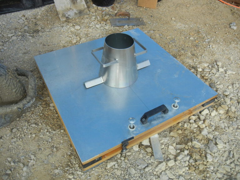 Das Bild zeigt den Aufbau eines Setzmaßversuchs zur Überwachung der Frischbetonkonsistenz. Auf einer Metallplatte mit Griffen und Halterungen steht ein metallischer Kegel (Setzmaßkegel), der zur Prüfung des Betonverhaltens vor dem Erhärten dient. Im Hintergrund ist eine Baustelle mit Kies und weiteren Baumaterialien zu erkennen.