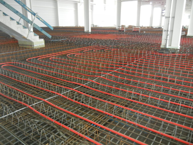 Auf dem Bild ist eine Baustelle mit einer großen Fläche aus bewehrtem Beton zu sehen. Der Boden ist mit einem Gitter aus Stahlbewehrung und roten Kunststoffrohren bedeckt, für eine Fußbodenheizung oder Kühlung. Metallstützen halten die Bewehrung in Position, und die Baustelle befindet sich innerhalb eines Gebäudes mit weißen Wänden und Säulen.
