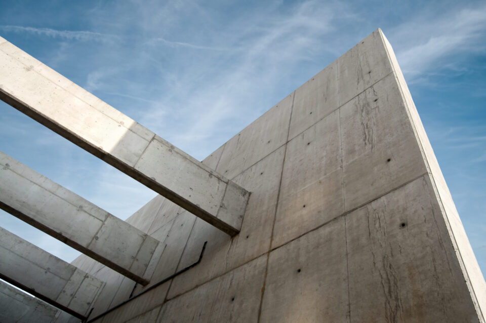 Das Bild zeigt eine Nahaufnahme eines Betonbauwerks mit großen, ebenen Flächen und auskragenden Trägern. Im Hintergrund ist der Himmel zu sehen. Die Struktur wirkt massiv und technisch.