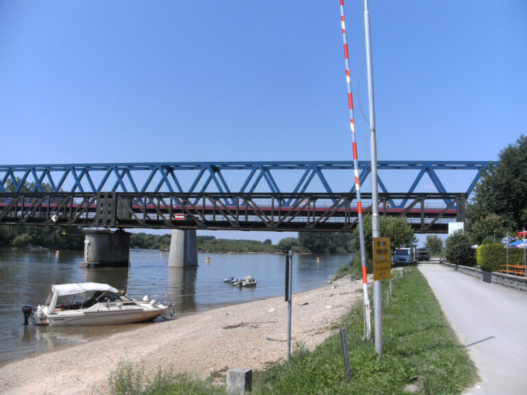 Das Bild zeigt eine blaue Fachwerkbrücke, die über einen Fluss führt und von Betonpfeilern getragen wird. Am Ufer liegen zwei Boote, eines im Vordergrund und ein kleineres weiter hinten, rechts ist eine Straße mit Schranke zu sehen.