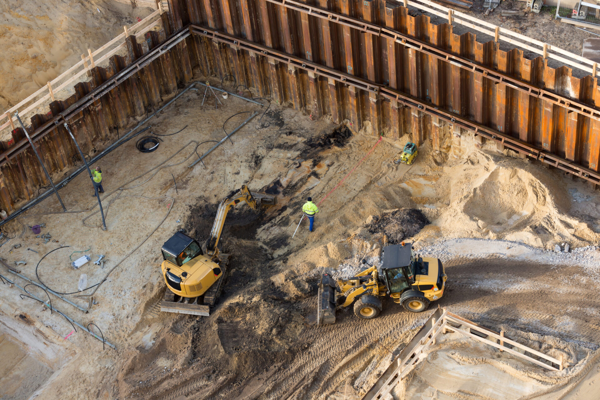 Das Bild zeigt eine Baustelle mit drei Arbeitern und zwei Baumaschinen. Der Bereich ist von großen Metallspundwänden umgeben, der Boden besteht überwiegend aus Sand und Erde. Ein Arbeiter bedient einen kleinen Bagger, ein weiterer nutzt ein Vermessungsgerät, und der dritte überwacht oder koordiniert die Arbeiten.