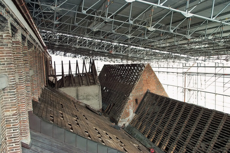 Das Bild zeigt ein altes, stark beschädigtes Gebäude mit mehreren schrägen Dächern, von denen viele Ziegel fehlen und Holzbalken freiliegen. Um das Gebäude herum ist ein Gerüst aufgebaut, was auf Renovierungs- oder Sicherungsarbeiten hinweist.