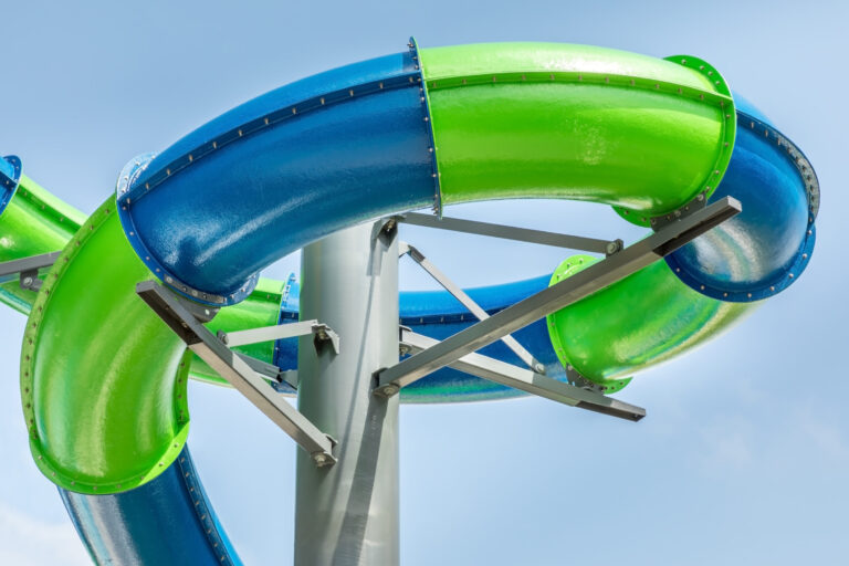 Das Bild zeigt eine Nahaufnahme einer Wasserrutsche mit zwei ineinander verschlungenen Röhren, eine blau und eine grün. Die Röhren sind glänzend und werden von Metallstreben an einem zentralen Mast gehalten. Im Hintergrund ist ein klarer, blauer Himmel zu sehen.