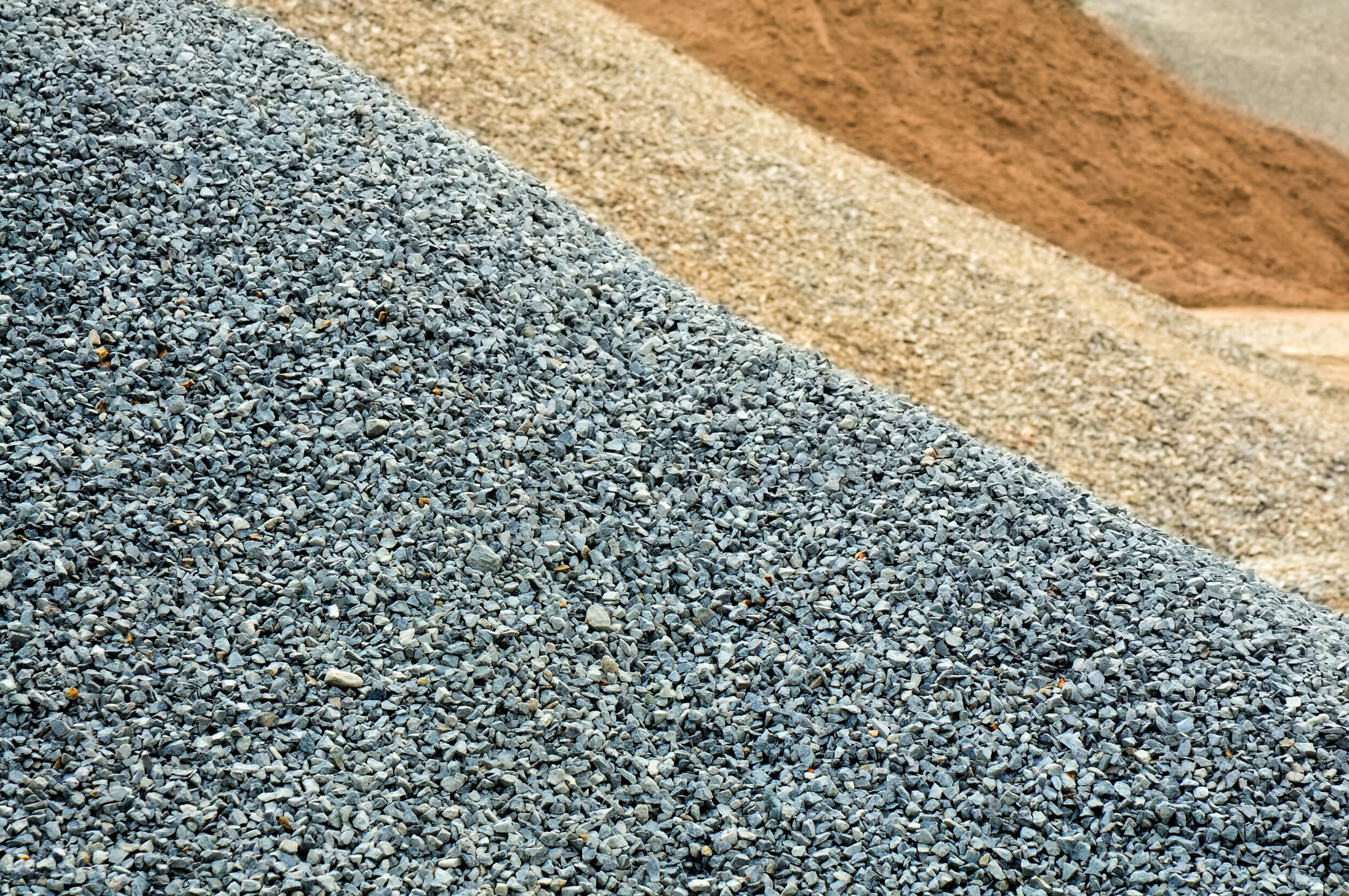 Das Bild zeigt mehrere Haufen verschiedener Baustoffe. Im Vordergrund liegt ein Haufen aus kleinem grauem Kies, dahinter befindet sich ein beiger Sandhaufen. Weiter hinten ist ein weiterer Haufen mit etwas größeren Steinen oder gebrochenem Gestein (Splitt) zu sehen.