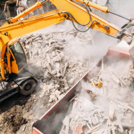 Das Bild zeigt eine Baustelle, auf der ein gelber Bagger Trümmer in einen großen Container lädt. Die Schaufel des Baggers ist mit Bauschutt gefüllt, darunter Holz- und Betonteile, wobei viel Staub aufgewirbelt wird. Die Umgebung deutet auf Abriss- oder Aufräumarbeiten hin.
