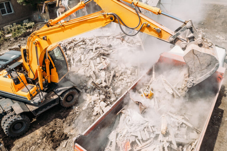 Das Bild zeigt eine Baustelle, auf der ein gelber Bagger Trümmer in einen großen Container lädt. Die Schaufel des Baggers ist mit Bauschutt gefüllt, darunter Holz- und Betonteile, wobei viel Staub aufgewirbelt wird. Die Umgebung deutet auf Abriss- oder Aufräumarbeiten hin.