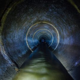 Das Bild zeigt einen dunklen unterirdischen Abwasserkanal mit einem runden Betontunnel. Schwaches Licht beleuchtet die raue Oberfläche der Betonwände. Die Szene wirkt industriell und funktional.