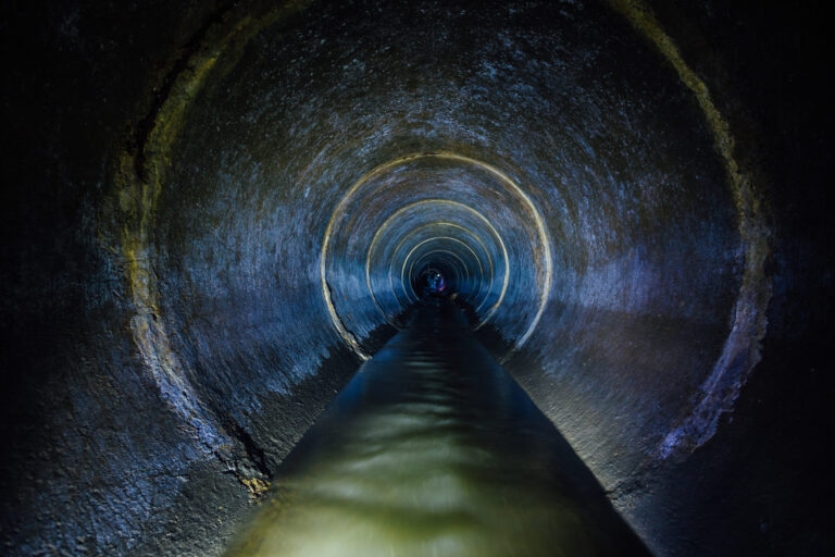 Das Bild zeigt einen dunklen unterirdischen Abwasserkanal mit einem runden Betontunnel. Schwaches Licht beleuchtet die raue Oberfläche der Betonwände. Die Szene wirkt industriell und funktional.