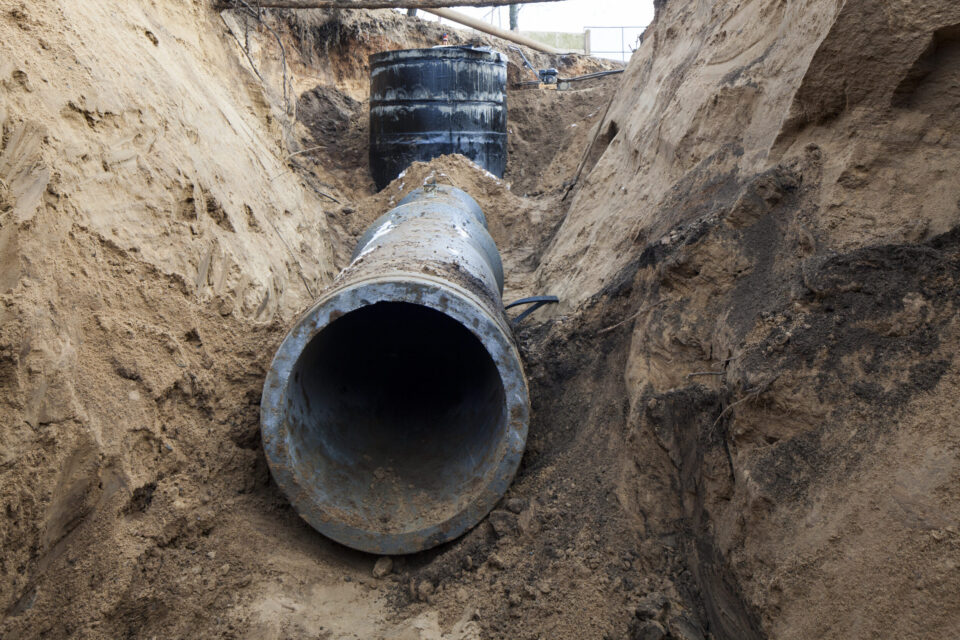 Das Bild zeigt ein großes, teilweise im Boden verlegtes Metallrohr in einem Graben. Der Graben ist in sandigen Boden gegraben, das Rohr gehört offenbar zu einem unterirdischen Infrastrukturprojekt. Im Hintergrund befindet sich eine weitere zylindrische Struktur, die mit dem Rohr verbunden zu sein scheint.