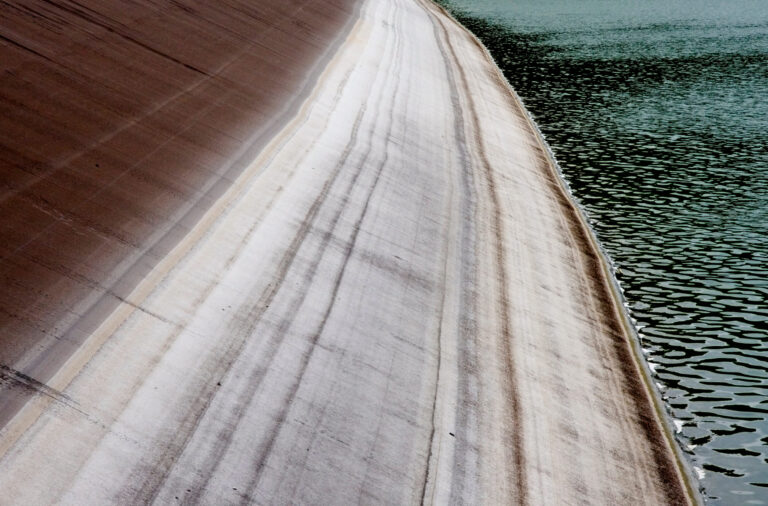 Das Bild zeigt eine geneigte Betonfläche mit deutlich sichtbaren horizontalen Linien, die auf unterschiedliche Wasserstände im Laufe der Zeit hinweisen. Rechts im Bild befindet sich ein Gewässer mit kleinen Wellen auf der Oberfläche. Die linke Seite der Betonfläche ist trocken und weist eine rötlich-braune Färbung auf.