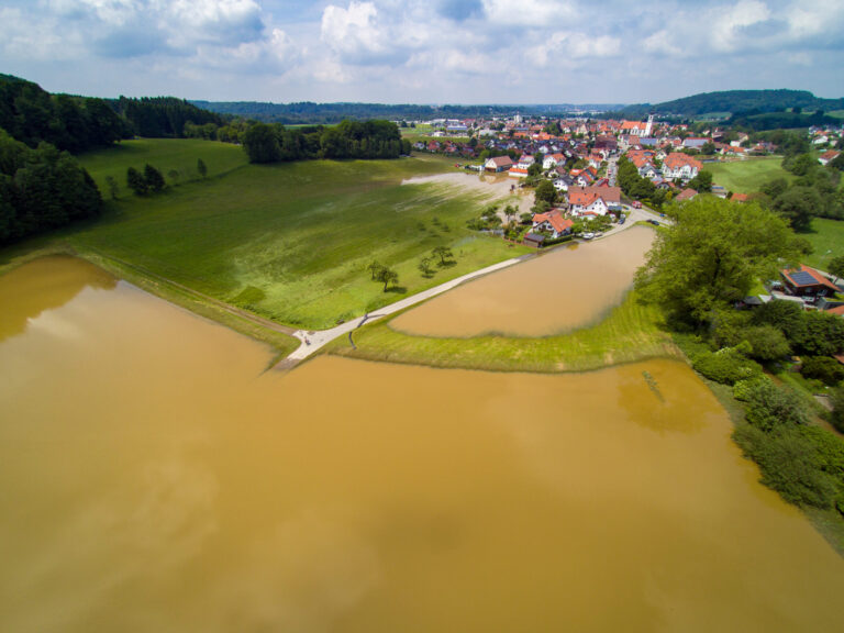 Das Bild zeigt eine Luftaufnahme einer Kleinstadt mit einem braun gefärbten Gewässer im Vordergrund, einer Überschwemmung. Umgeben ist die Stadt von grünen Feldern, Baumgruppen und sanften Hügeln im Hintergrund die teilweise überflutet wurden.