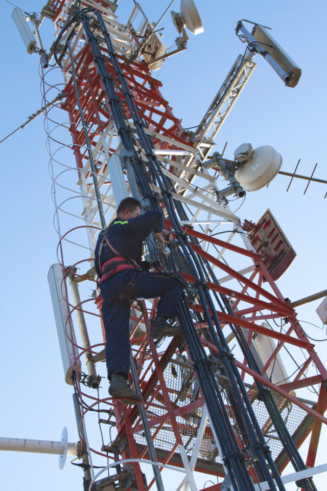 Das Bild zeigt einen Arbeiter, der einen hohen Kommunikationsturm erklimmt. Der Turm besteht aus rot-weiß gestrichenen Metallstrukturen und ist mit verschiedenen Antennen und Satellitenschüsseln ausgestattet. Der Arbeiter trägt Sicherheitsausrüstung einschließlich eines Gurts und führt Wartungsarbeiten durch.