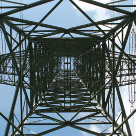 Das Bild zeigt den Blick von unten auf einen Hochspannungsmast, wobei die komplexe Gitterstruktur aus Metallträgern und Stützen sichtbar ist. Durch die geometrischen Muster der Konstruktion ist der Himmel mit einigen Wolken zu erkennen. Entlang beider Seiten des Masts verlaufen mehrere Stromleitungen senkrecht nach oben.