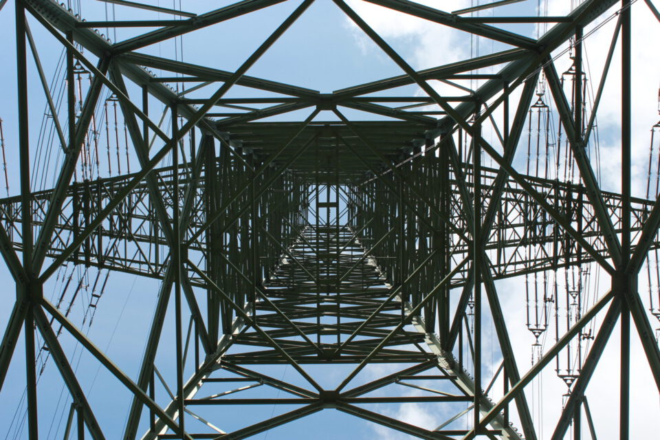 Das Bild zeigt den Blick von unten auf einen Hochspannungsmast, wobei die komplexe Gitterstruktur aus Metallträgern und Stützen sichtbar ist. Durch die geometrischen Muster der Konstruktion ist der Himmel mit einigen Wolken zu erkennen. Entlang beider Seiten des Masts verlaufen mehrere Stromleitungen senkrecht nach oben.