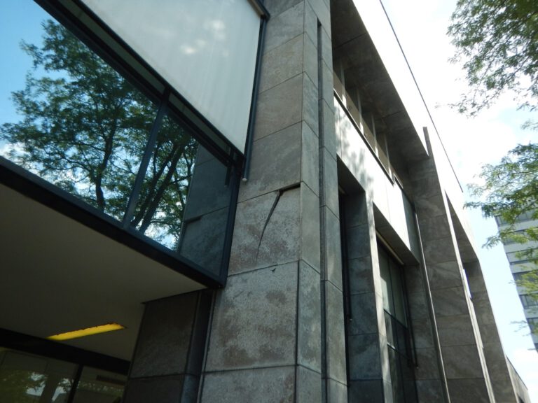 Das Bild zeigt die Außenansicht eines modernen Gebäudes mit großen Fenstern und einer Fassade aus rechteckigen Steintafeln. In den Fenstern spiegeln sich Bäume und der Himmel, was auf eine grüne Umgebung hinweist.