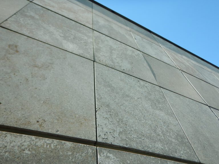 Das Bild zeigt eine Nahaufnahme der Außenwand eines Gebäudes, bestehend aus großen rechteckigen Beton- oder Steinplatten. Die Platten sind in einem Rastermuster angeordnet und weisen sichtbare Fugen auf. Die Oberfläche wirkt verwittert und leicht verfärbt, was auf längere Witterungseinflüsse hinweist.