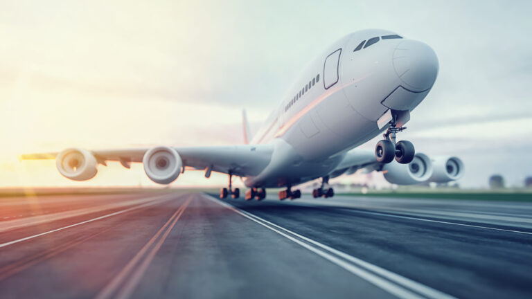 Ein großes Verkehrsflugzeug hebt von einer Asphaltpiste ab. Das vordere Fahrwerk ist bereits in der Luft, während das hintere noch den Boden berührt. Im Hintergrund ist ein klarer Himmel mit einigen Wolken zu sehen, beleuchtet von Sonnenlicht von der linken Seite.
