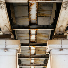 Das Bild zeigt die Unterseite einer Brücke oder eines Überführungsbauwerks, das von massiven Betonsäulen getragen wird. Die Konstruktion enthält Metallträger mit sichtbaren Rostspuren, was auf Witterungseinflüsse hinweist. Die Betonsäulen wirken robust und tragen die darüberliegende Struktur.