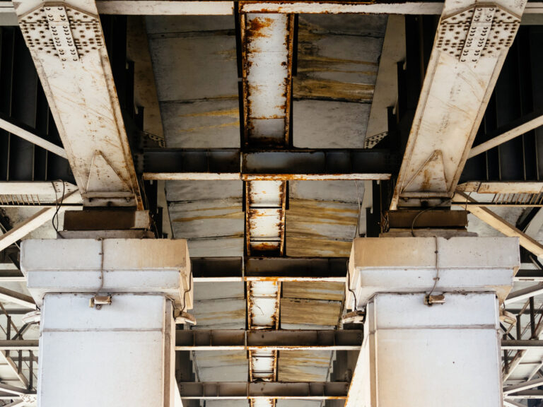Das Bild zeigt die Unterseite einer Brücke oder eines Überführungsbauwerks, das von massiven Betonsäulen getragen wird. Die Konstruktion enthält Metallträger mit sichtbaren Rostspuren, was auf Witterungseinflüsse hinweist. Die Betonsäulen wirken robust und tragen die darüberliegende Struktur.