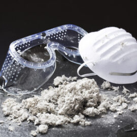 Das Bild zeigt eine Schutzbrille und eine weiße Staubmaske, die auf einer dunklen Oberfläche liegen. Davor befindet sich ein Haufen faseriges Material, das wie Asbest oder ein ähnlicher Stoff aussieht. Der schwarze Hintergrund hebt die Gegenstände im Vordergrund deutlich hervor.