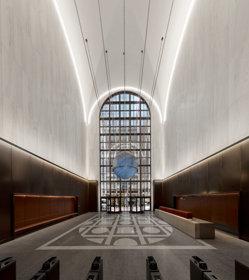 Das Bild zeigt eine moderne, großzügige Eingangshalle mit hohen Decken und einem großen Rundbogenfenster am Ende. Die Wände bestehen aus hellem Marmor, in der Mitte des Bodens befindet sich ein geometrisches Muster. Vor dem Fenster schwebt eine blaue Kugelskulptur, und an der Decke verlaufen LED-Lichtstreifen. Am Eingang sind mehrere Zugangsschranken zu sehen.