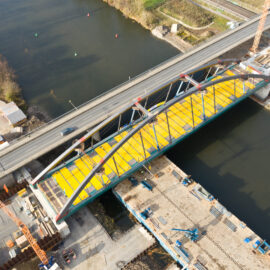 Luftaufnahme einer Brücke im Bau mit gelbem Deck, die von zwei großen Bögen getragen wird und über einen Fluss führt. Auf beiden Seiten der Brücke befinden sich Baustellen mit Kränen und Baugeräten, ein Auto fährt über die Brücke.